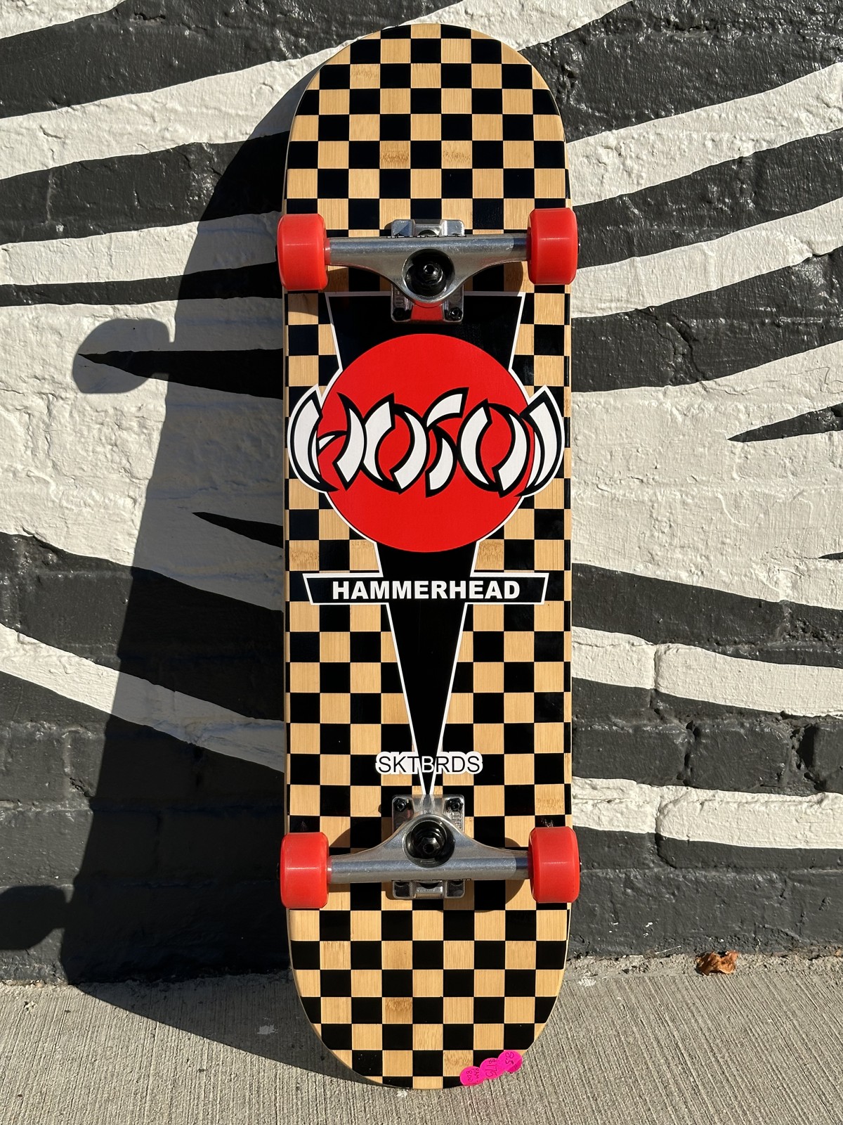 Hosoi Hammerhead complete skateboard with checkerboard deck and red wheels leaning against a striped wall