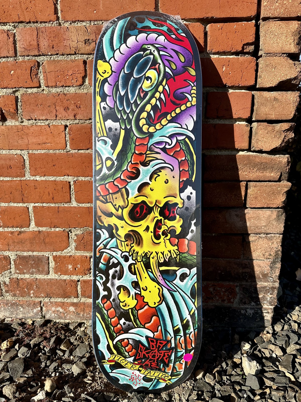 Skateboard deck with tattoo-style snake and yellow skull art and a Tiger Tattoo banner, leaning against a brick wall