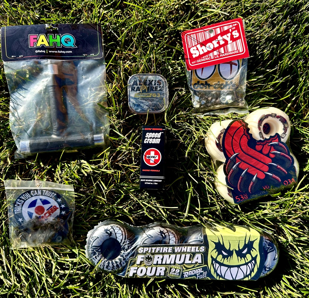 Skate hardware and accessories laid out on grass: skate tool, bolts, bearings, speed cream, wheels, and hardware